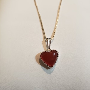 May include: A silver chain necklace with a red heart pendant. The heart is set in a silver frame with a scalloped edge.