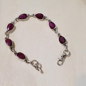 May include: A silver bracelet featuring teardrop-shaped, deep red gemstones. The gemstones are linked together with silver connectors and a clasp closure. The bracelet is displayed on a light-colored surface.