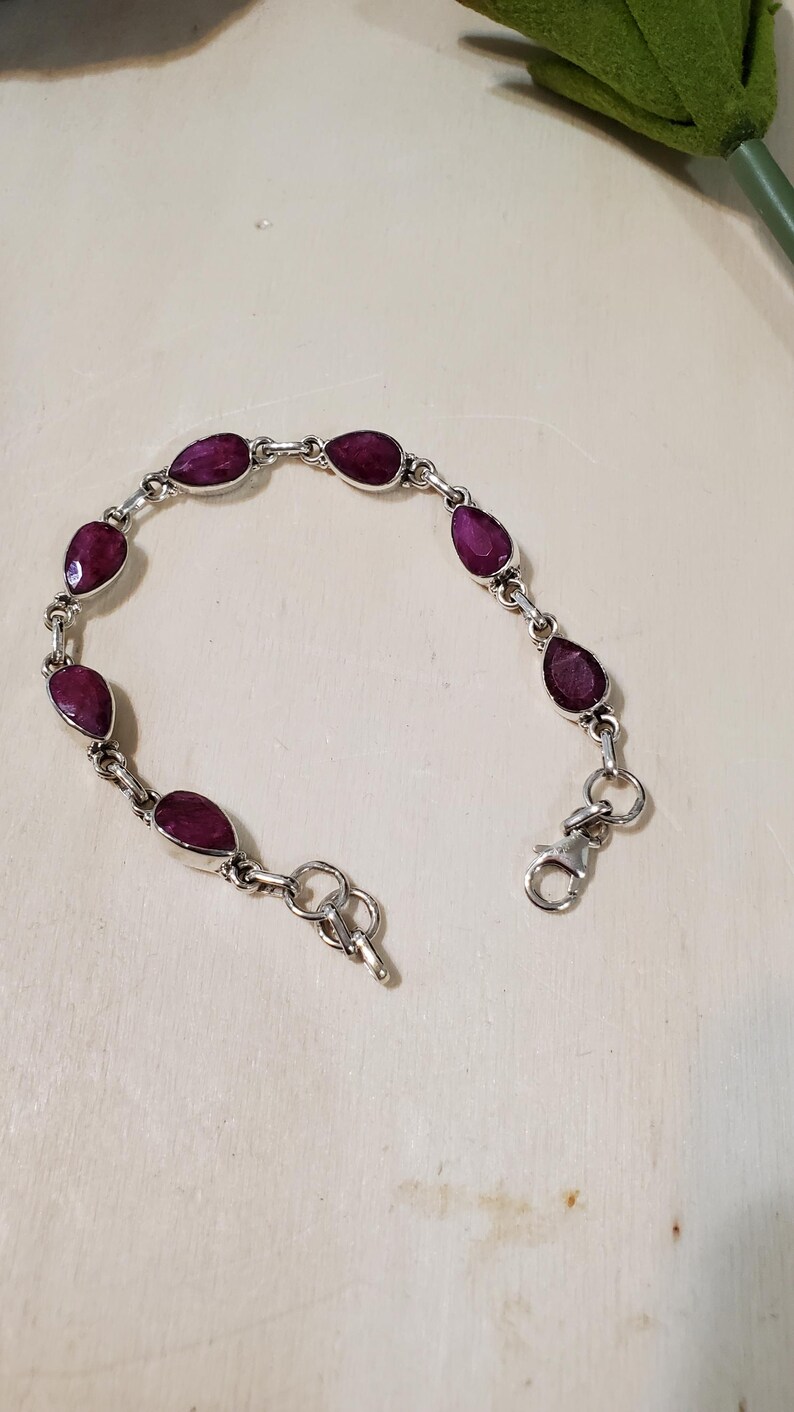 May include: A silver bracelet featuring teardrop-shaped, deep red gemstones. The gemstones are linked together with silver connectors and a clasp closure. The bracelet is displayed on a light-colored surface.