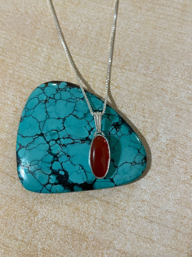 May include: A silver chain necklace with a red carnelian gemstone pendant. The pendant is oval shaped and set in a silver bezel.