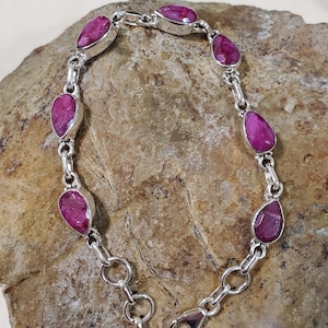 May include: A silver bracelet featuring teardrop-shaped, vibrant pink gemstones. The gemstones are set in individual silver bezels and linked together with silver chain. The bracelet is displayed on a textured, brown stone surface.