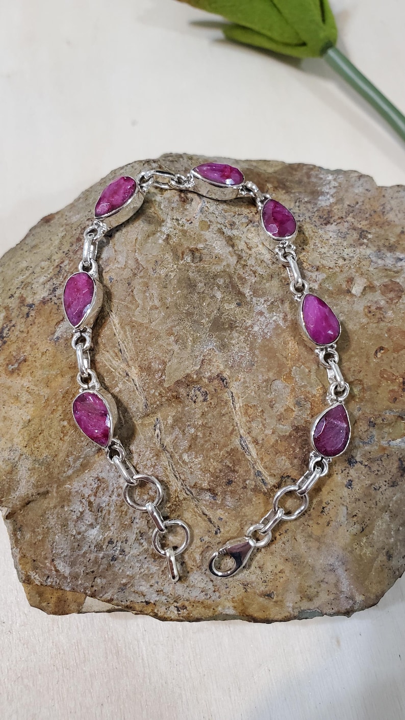 May include: A silver bracelet featuring teardrop-shaped, vibrant pink gemstones. The gemstones are set in individual silver bezels and linked together with silver chain. The bracelet is displayed on a textured, brown stone surface.