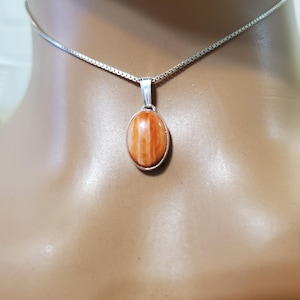May include: A silver necklace with an oval pendant. The pendant features an orange and white stone set in a silver bezel. The necklace is displayed on a mannequin.