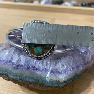 May include: A silver bracelet with a turquoise stone set in a silver bezel. The stone is approximately 2.5 cm in diameter. The bracelet is on a purple amethyst geode.