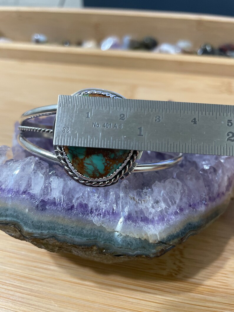 May include: A silver bracelet with a turquoise stone set in a silver bezel. The stone is approximately 2.5 cm in diameter. The bracelet is on a purple amethyst geode.
