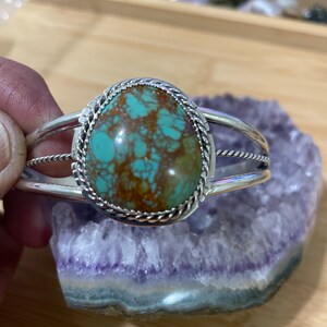 May include: A silver bracelet featuring a turquoise stone set within a rope-like border. The stone has a green and brown marbled pattern.