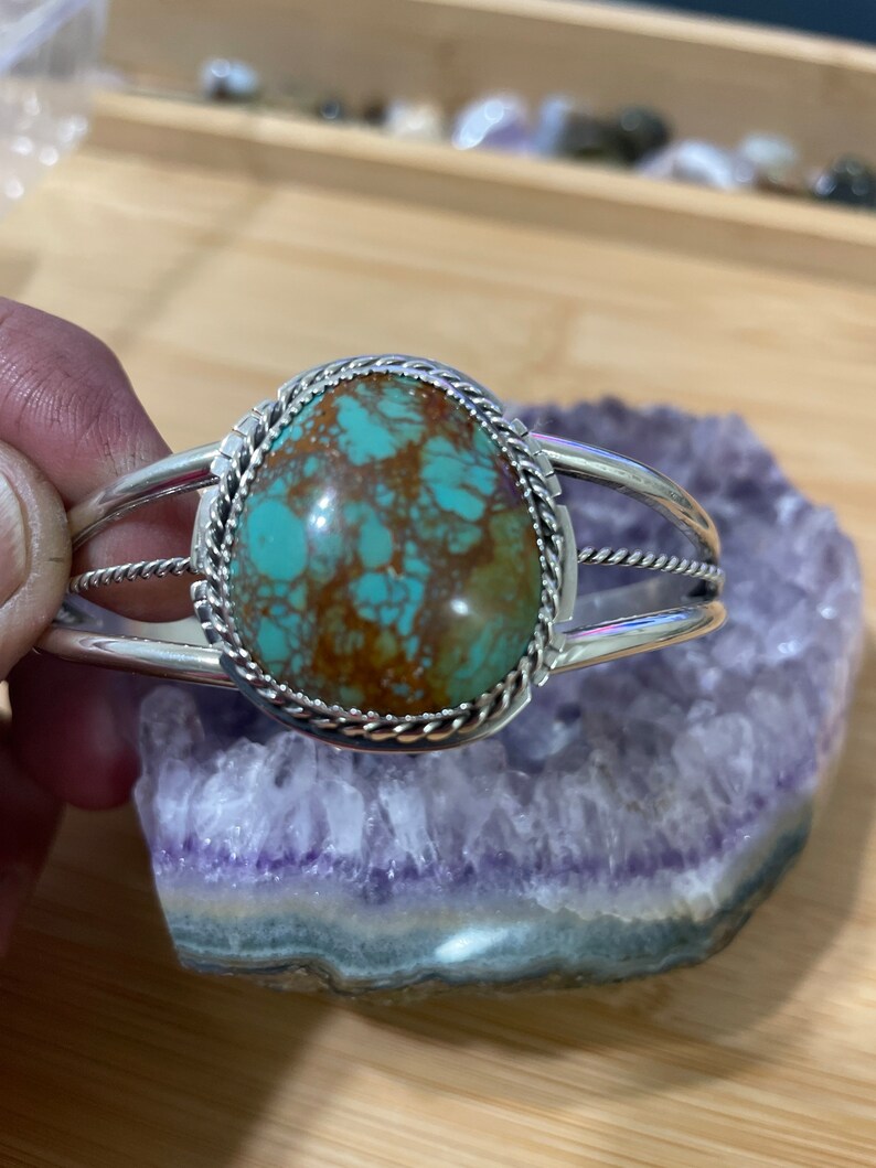 May include: A silver bracelet featuring a turquoise stone set within a rope-like border. The stone has a green and brown marbled pattern.