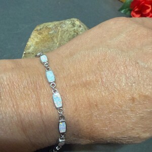 May include: A silver bracelet featuring rectangular opal stones. The bracelet is composed of small, interconnected links. The opals have a milky white and iridescent appearance. The bracelet is worn on a wrist.