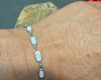 May include: A silver bracelet featuring rectangular opal stones. The bracelet is composed of small, interconnected links. The opals have a milky white and iridescent appearance. The bracelet is worn on a wrist.