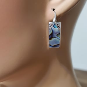 May include: Silver rectangular earrings with a vibrant abalone shell inlay. The earrings have a decorative filigree border and a simple hook closure. The abalone shell displays iridescent blues, greens, and purples.