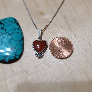 May include: A silver chain necklace with a heart-shaped pendant featuring a red carnelian stone. The pendant is surrounded by a silver border and has small silver beads at the bottom. The necklace is shown next to a US penny for size comparison.