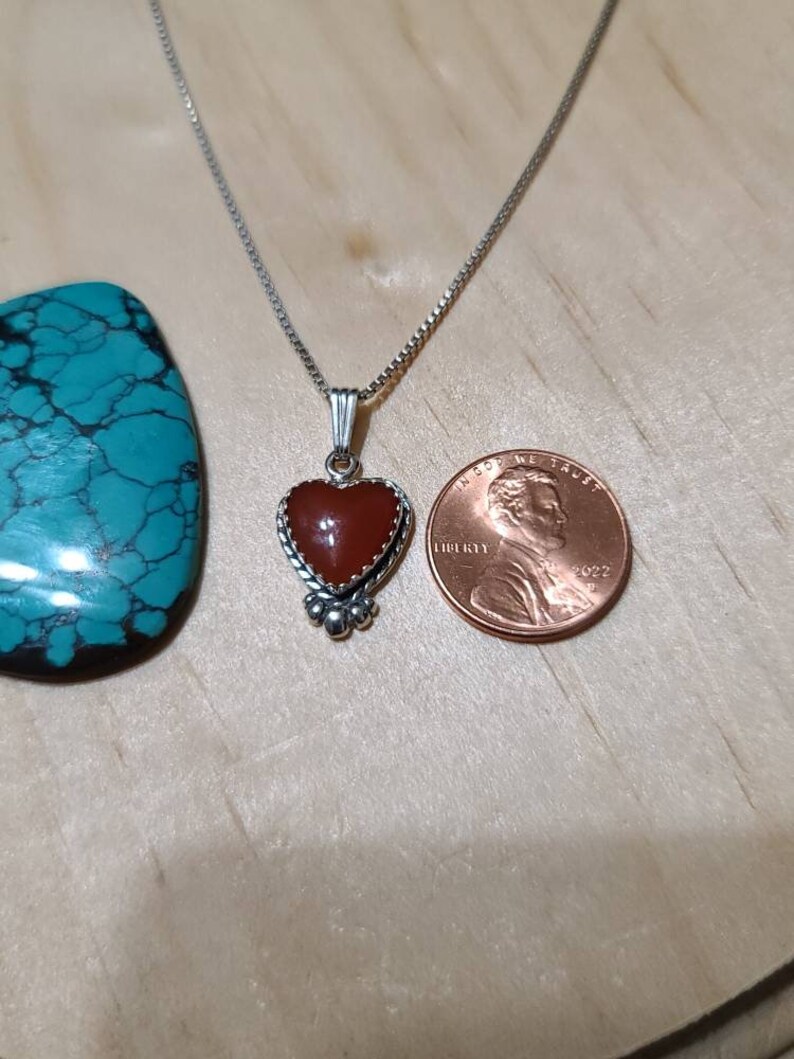 May include: A silver chain necklace with a heart-shaped pendant featuring a red carnelian stone. The pendant is surrounded by a silver border and has small silver beads at the bottom. The necklace is shown next to a US penny for size comparison.