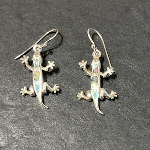 May include: A pair of silver earrings featuring a detailed lizard design with iridescent abalone shell accents.