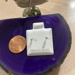 May include: A pair of sterling silver stud earrings with small clear stones. The earrings are displayed on a white card with the words "Sterling Silver" printed on it. The card is sitting on a purple and white agate slice.