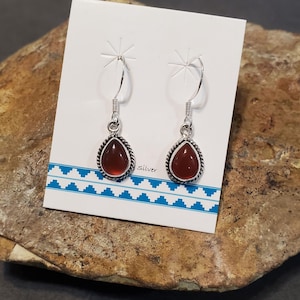 May include: A pair of teardrop-shaped earrings featuring smooth, deep red stones set in twisted silver-tone metal frames. The earrings hang from silver-tone hooks and are displayed on a white card with a blue geometric pattern and the word Silver.