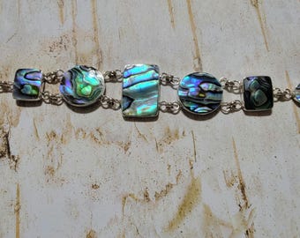 May include: A silver bracelet with iridescent abalone shell accents. The bracelet features a mix of round and rectangular shapes.