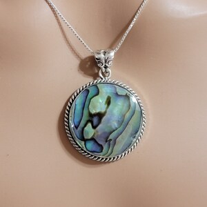 May include: A silver chain necklace with a round abalone shell pendant. The pendant has a silver rope border and a blue, green, and purple iridescent shell.