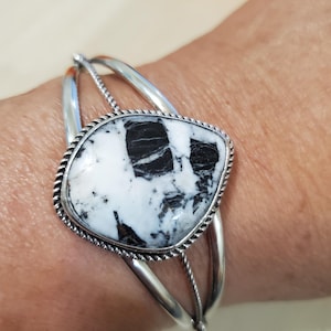 May include: A silver cuff bracelet featuring a large, irregularly shaped white stone with black veining. The stone is framed by a twisted rope-like border. The bracelet has a simple, open design with two curved bands.
