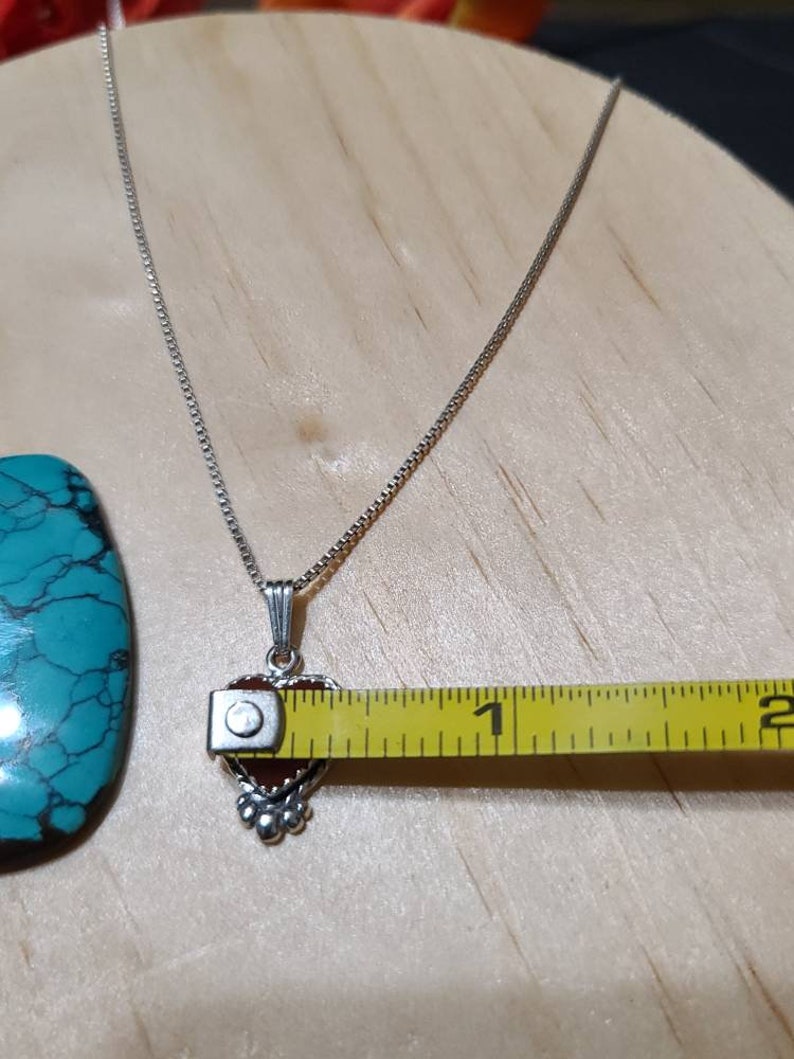 May include: A silver chain necklace with a heart-shaped pendant featuring a red gemstone. The pendant is approximately 1 inch in length.