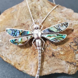 May include: A silver dragonfly necklace with iridescent abalone shell wings. The necklace is on a rock background.