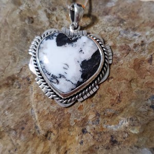 May include: A heart-shaped pendant necklace with a white and black stone set in a silver-toned metal frame. The pendant has a decorative border and hangs from a delicate chain. The necklace is displayed on a textured, brown surface.