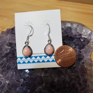 May include: A pair of sterling silver earrings with teardrop-shaped, light pink stones. The stones are set in a silver bezel with a twisted rope design. The earrings hang from silver fish hook ear wires. A penny is next to the earrings.