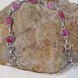 May include: A silver bracelet featuring oval-shaped, vibrant pink gemstones. The bracelet is composed of interconnected links, with each gemstone set within a decorative bezel. The clasp is visible at the end of the bracelet. The bracelet is displayed on a textured, light brown surface.