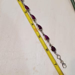 May include: A silver bracelet with teardrop-shaped, deep red gemstones. The bracelet is measured with a yellow tape measure, showing a length of approximately 7 inches. The gemstones are set in silver bezels.