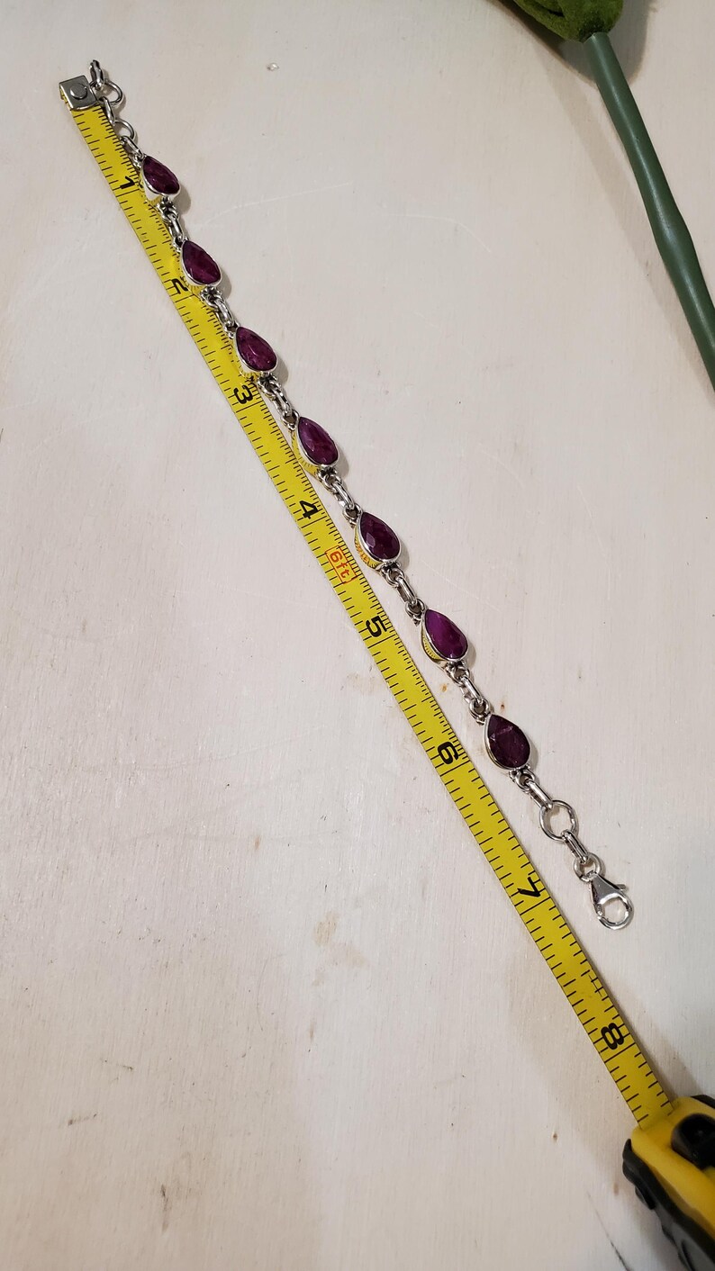 May include: A silver bracelet with teardrop-shaped, deep red gemstones. The bracelet is measured with a yellow tape measure, showing a length of approximately 7 inches. The gemstones are set in silver bezels.