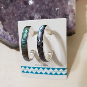 May include: A pair of silver hoop earrings with a blue and green abalone shell inlay. The earrings are displayed on a white card with the text "sterling silver".