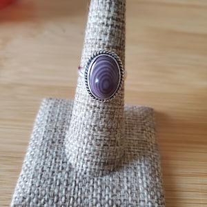 May include: A silver ring with an oval, purple stone. The stone has a striped pattern and is set in a silver bezel with a twisted rope design. The ring is displayed on a beige ring holder.