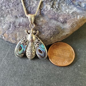 May include: A silver-toned bee pendant necklace with iridescent abalone wings. The bee pendant is detailed with intricate designs and hangs from a delicate chain. A US one-cent coin is next to the pendant for size reference.