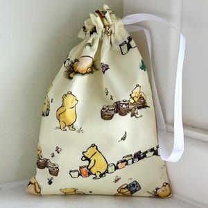 May include: A drawstring bag featuring a repeating pattern of a yellow bear with honey pots and butterflies on a cream background. The bag has a white ribbon drawstring closure and is made of fabric.