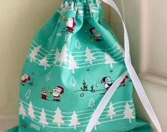 May include: A teal drawstring gift bag with a repeating pattern of white Christmas trees and Santa Claus figures. The bag has a white ribbon drawstring and the words "Jingle" are visible on the fabric.