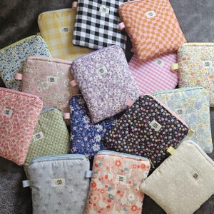 Puede incluir: Una colección de estuches acolchados con diversos diseños florales, de cuadros y estampados. Los estuches tienen cierres de cremallera y pequeñas etiquetas de tela. Los colores incluyen rosa, azul, amarillo, negro y blanco. Los estuches son probablemente para llevar objetos pequeños.