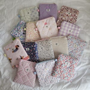 Può includere: Una collezione di pochette in tessuto trapuntato con vari disegni floreali e fantasia. Le pochette presentano una varietà di colori, tra cui rosa, blu e lavanda. Alcune pochette hanno un fiocco o una chiusura a cerniera. Le pochette sono disposte su una superficie bianca.