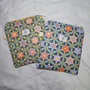 Op de afbeelding: Twee rechthoekige etuis met ritssluiting en een geometrisch rasterpatroon met kleurrijke bloemmotieven. De ene heeft een donkergroene achtergrond met een gele rits, de andere een donkerblauwe achtergrond met een lichtblauwe rits. Elk heeft een wit label met still with you.