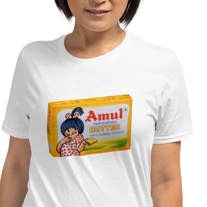 Puede incluir: Una camiseta blanca con un estampado amarillo y rojo de una niña de dibujos animados sosteniendo una barra de mantequilla. El estampado dice "Amul Pasteurised Butter utterly buttery delicious".
