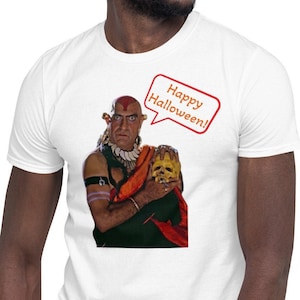 May include: A white t-shirt with a graphic of a man in a red and orange costume holding a golden mask. The text "Happy Halloween!" is in a speech bubble above the man.