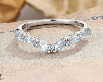 May include: A delicate silver band with a curved row of small, clear gemstones. The ring is set on a textured, cream-colored surface.