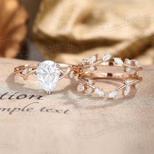 May include: A pear-shaped diamond engagement ring with a delicate, twisted band and a matching leaf-shaped wedding band. Both rings are set in rose gold and feature small diamonds.