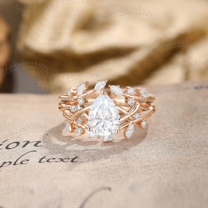 May include: A pear-shaped diamond engagement ring set in rose gold with a delicate floral design. The ring features a pear-shaped center stone surrounded by smaller diamonds and intricate floral details.