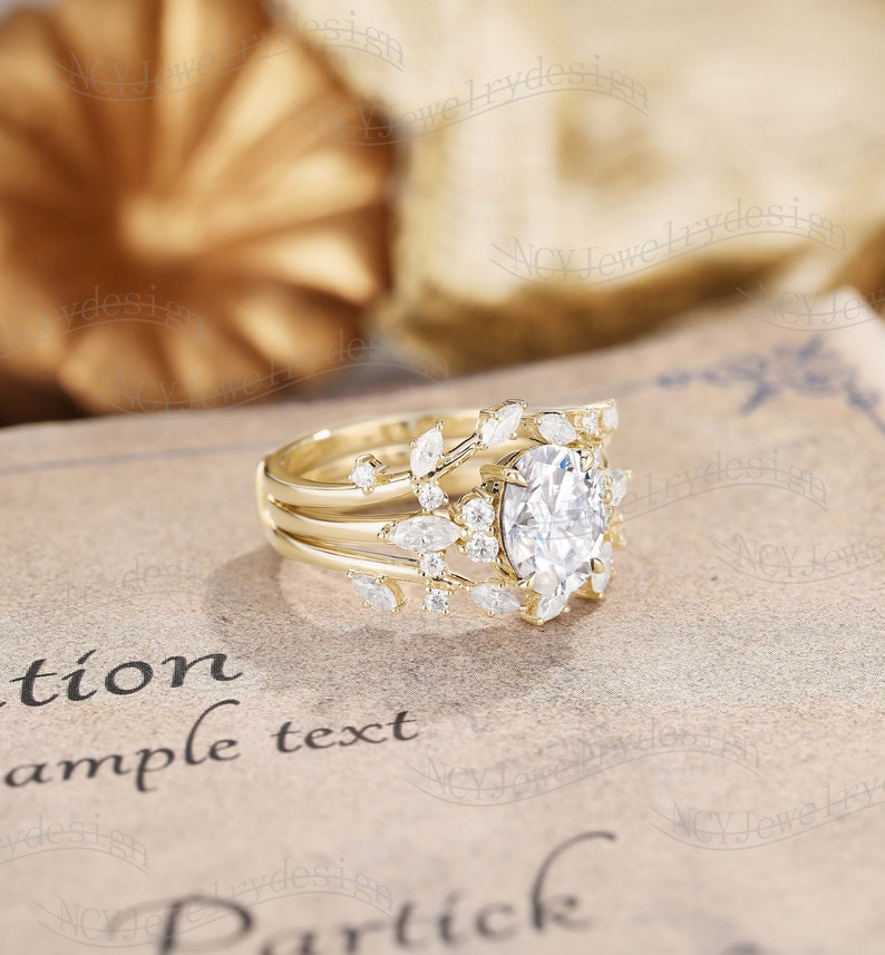 Op de afbeelding: Een gouden ring met een grote ovale middelste steen omringd door kleinere stenen. De ring is bezet met een delicaat bladontwerp en heeft een band met drie rijen kleinere stenen.