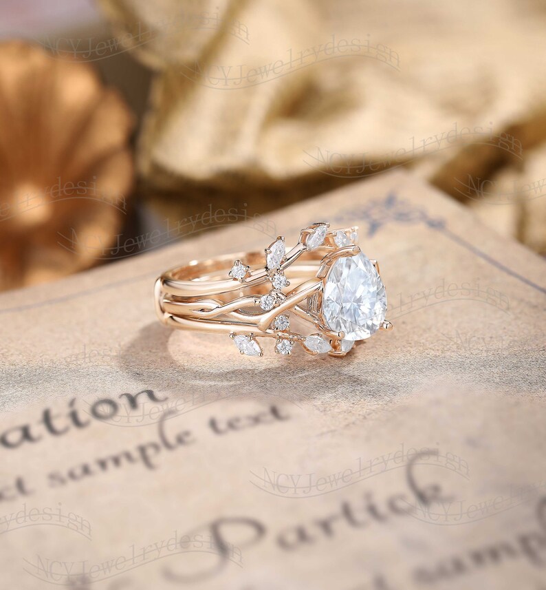 May include: A pear-shaped diamond engagement ring set in rose gold with a delicate, vine-like band adorned with smaller diamonds. The ring is set on a vintage-looking book with the text "Partick" visible.