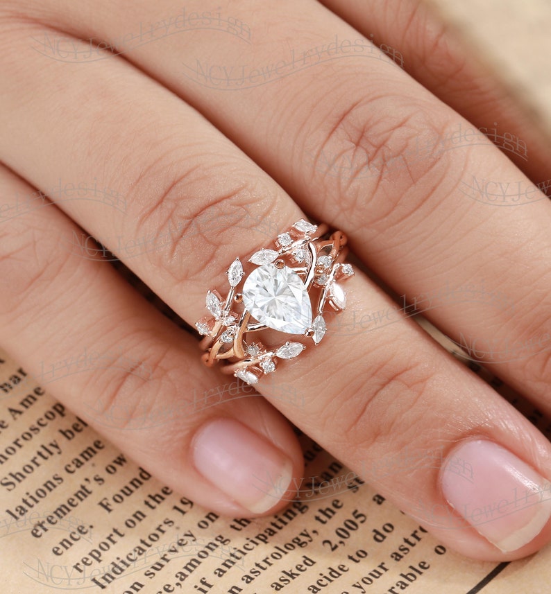 May include: A pear-shaped diamond engagement ring set in rose gold with a delicate leaf design. The ring features a pear-shaped center stone surrounded by smaller diamonds and leaves.