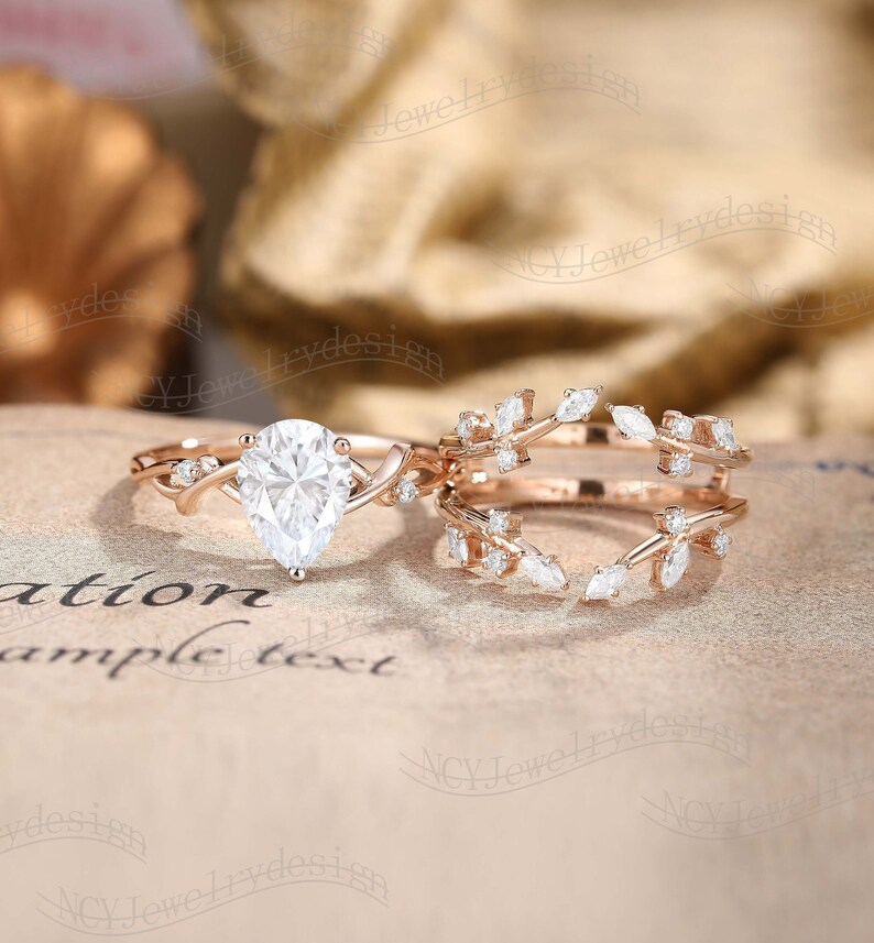May include: A pear-shaped diamond engagement ring with a delicate, twisted band and a matching leaf-shaped wedding band. Both rings are set in rose gold and feature small diamonds.