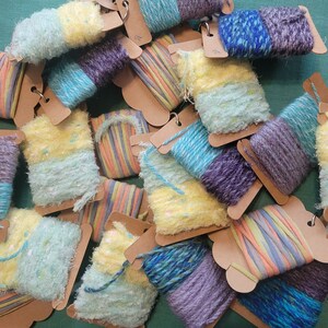May include: Assortment of colorful yarn skeins wrapped on cardboard spools. The yarn is a mix of textures, including fuzzy, smooth, and striped. The colors include yellow, blue, purple, and green.