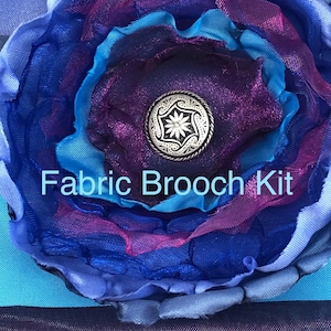 Puede incluir: Un kit de broche de tela con capas de pétalos de tela azules, morados y turquesas. El centro presenta un botón plateado con un diseño floral. El texto "Fabric Brooch Kit" se muestra en la imagen.