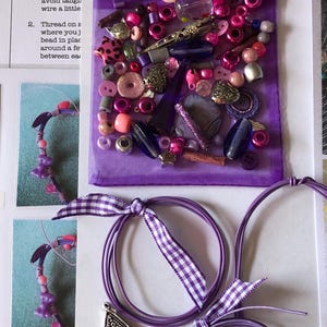 May include: A purple organza drawstring bag filled with an assortment of beads in shades of pink, purple, and silver, alongside two purple cord loops tied with purple and white gingham ribbon and a silver-toned triangular metal charm pendant.
