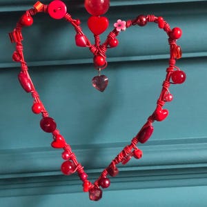May include: A heart-shaped ornament constructed from red wire, adorned with an assortment of red beads, buttons, and small heart-shaped charms. A single small pink flower bead is attached to the upper right side of the wire frame.